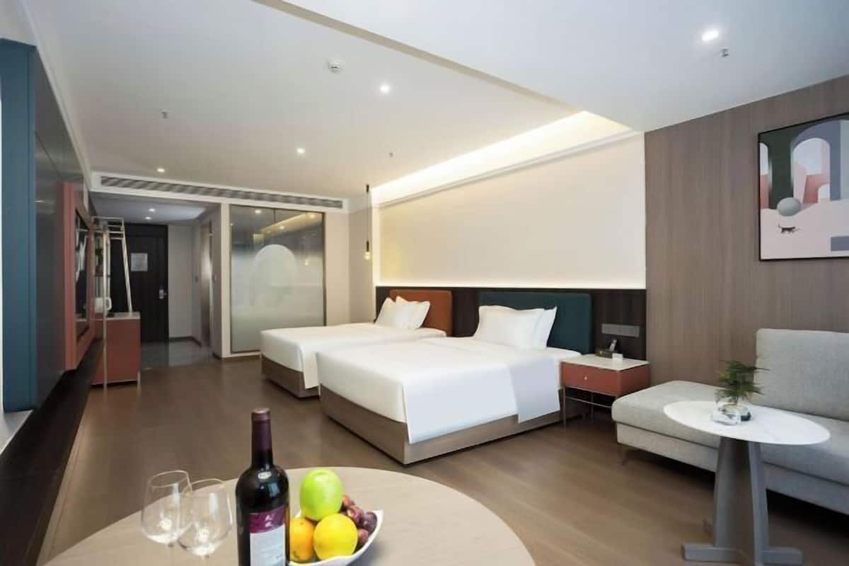 A hotel room with two beds, a sofa, a small table with a bottle and fruit, and a painting on the wall.