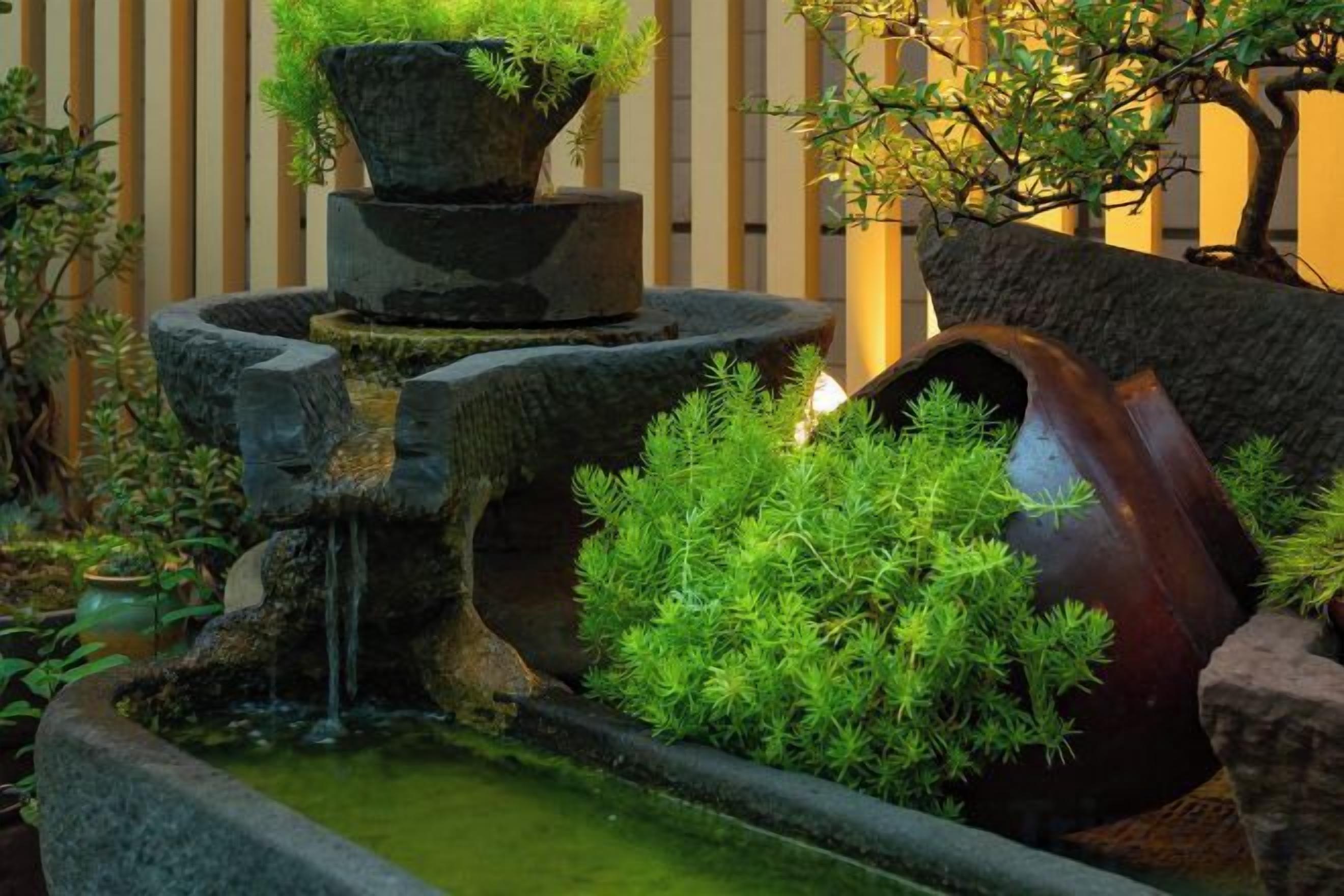Un jardín japonés tradicional con una fuente de agua de piedra, un puente de madera y abundante vegetación.