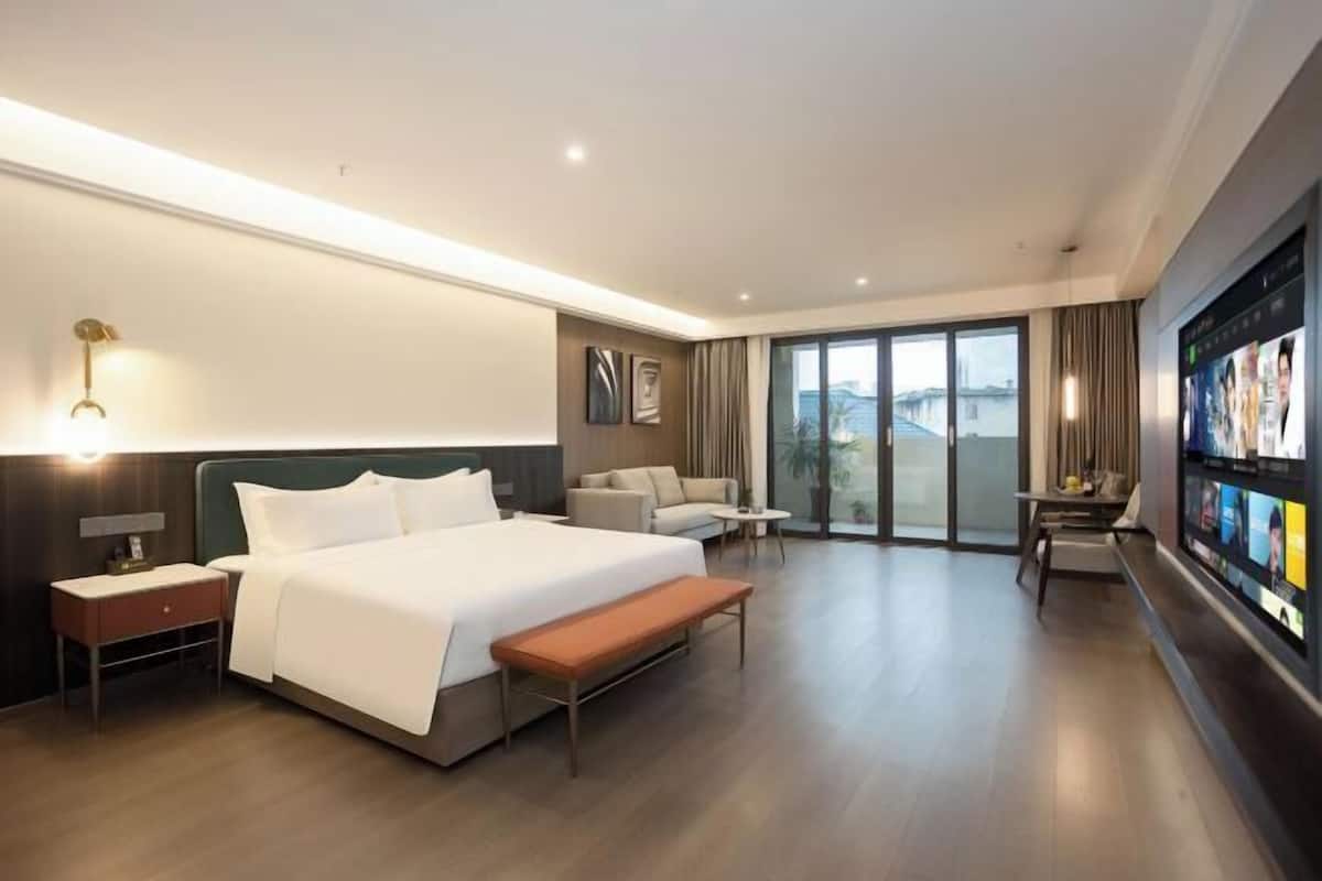 A modern hotel room with a large bed, a bench, a sofa, a TV, and a balcony with a view.