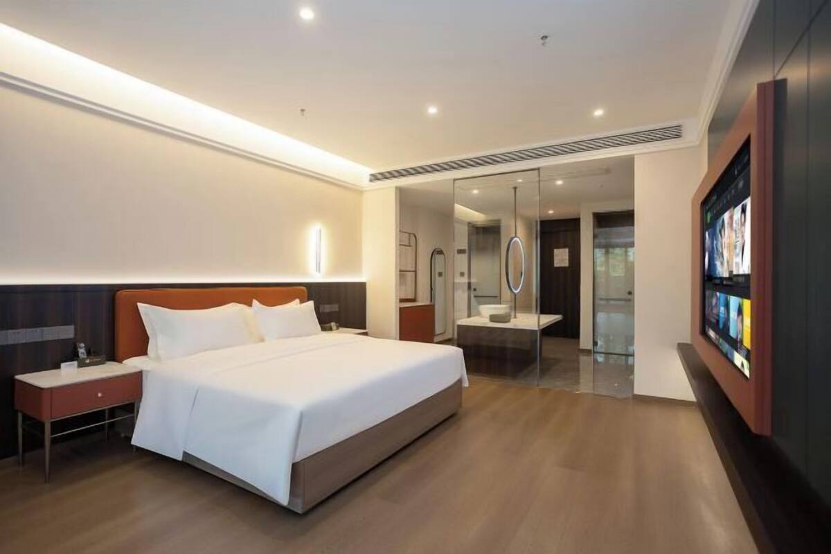 A modern hotel room with a large bed, bedside tables, a TV, and a glass-enclosed bathroom.