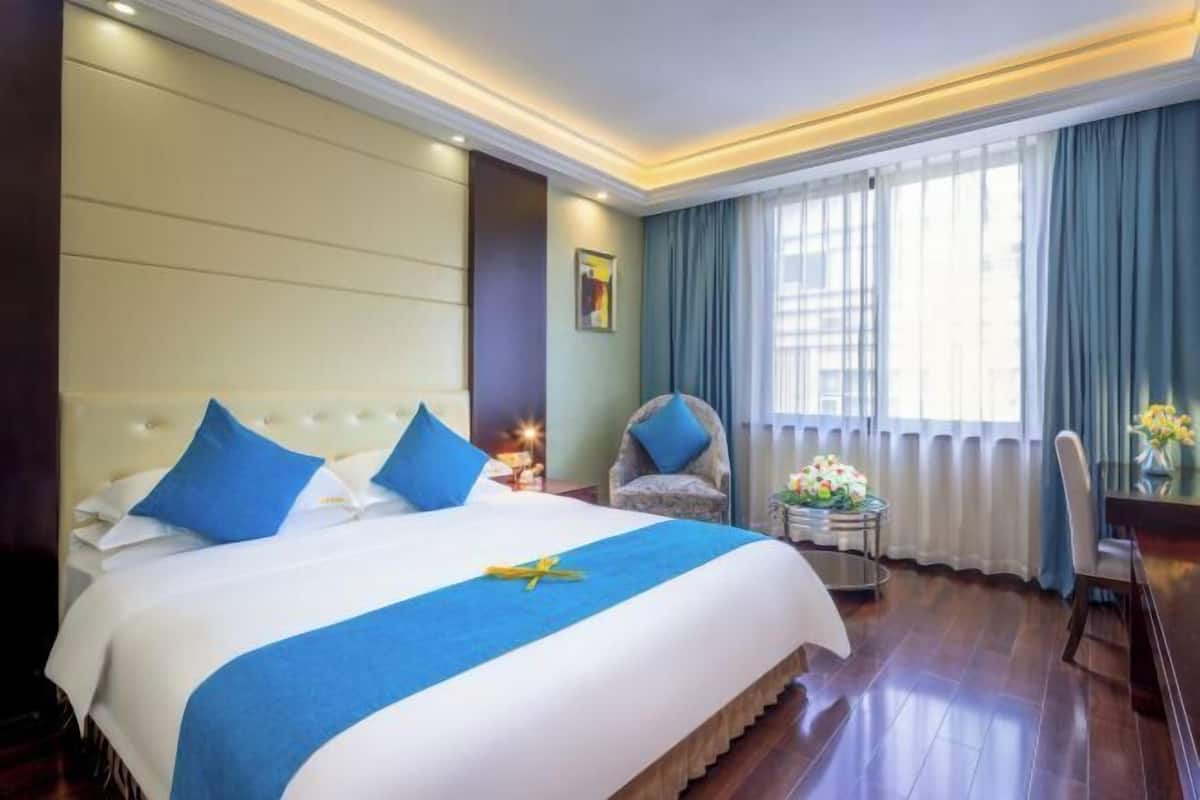 A hotel room with a large bed, a chair, a small table with flowers, and a window with curtains.