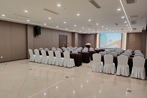 Un salón de conferencias con hileras de sillas, una pantalla de proyector y una mesa para presentaciones.