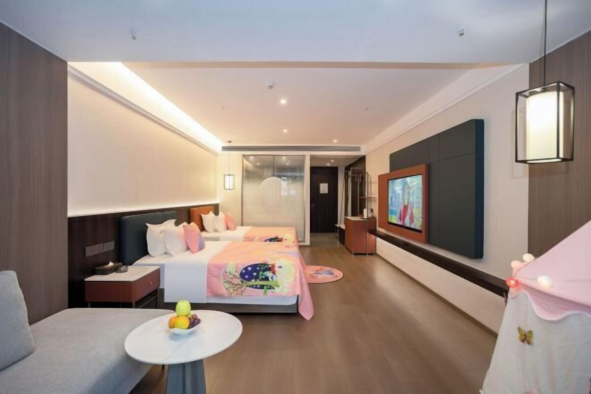 A modern hotel room with a bed, a TV, a small table with fruit, and a chair.