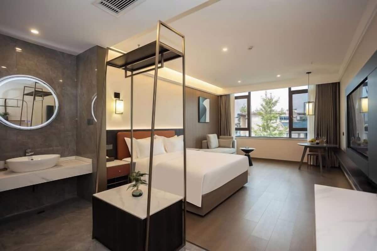 A hotel room with a large bed, a round mirror, a bedside table, a bathroom with a sink, and a view of the city through large windows.