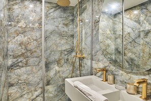 Ein Badezimmer mit Marmorwänden, einer gläsernen Dusche und einem weißen Waschbecken mit goldfarbenen Armaturen.