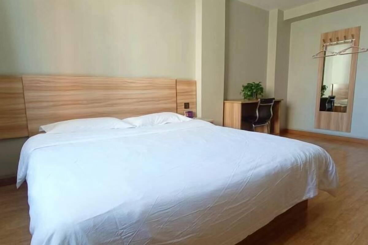 A bedroom with a large bed, a wooden headboard, a desk with a chair, a mirror, and a plant.