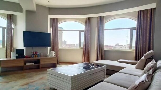 Una sala de estar moderna con una televisión de pantalla plana, un sofá, una mesa de centro y ventanas grandes con cortinas.