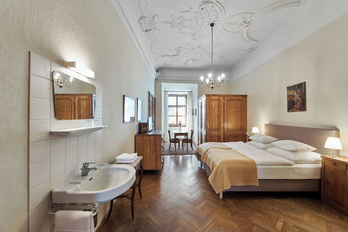 A hotel room with a bed, bedside table, lamp, wooden wardrobe, mirror, and a bathroom with a sink and chair.