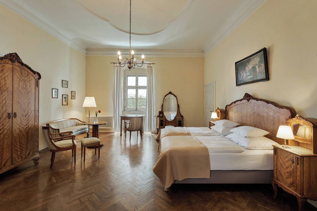 A hotel room with a large bed, wooden furniture, a chandelier, and a window with curtains.