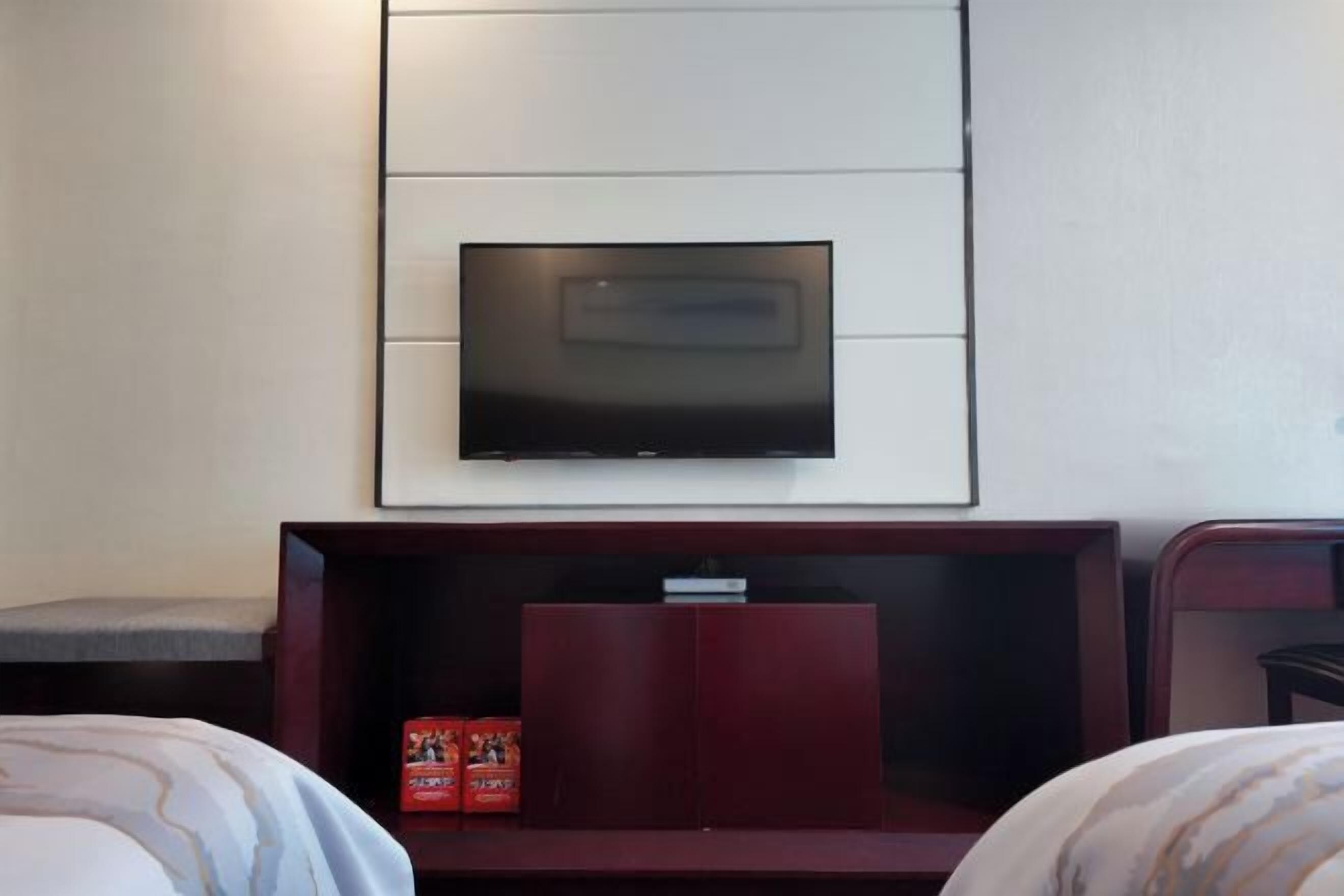 A hotel room with a flat-screen TV mounted on the wall, a wooden entertainment unit below it, and two beds with patterned bedding.