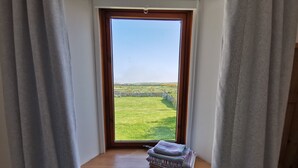 A window with a view of a green field and a stone wall. A wooden shelf with books and a folded towel is in front of the window. - Cosy Highland retreat with countryside and sea views. (John o' Groats)