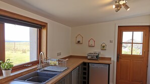 A compact kitchen with a sink, a dishwasher, and a toaster. There is a window providing a view of the outdoors, and a door leading to another room. - Cosy Highland retreat with countryside and sea views. (John o' Groats)