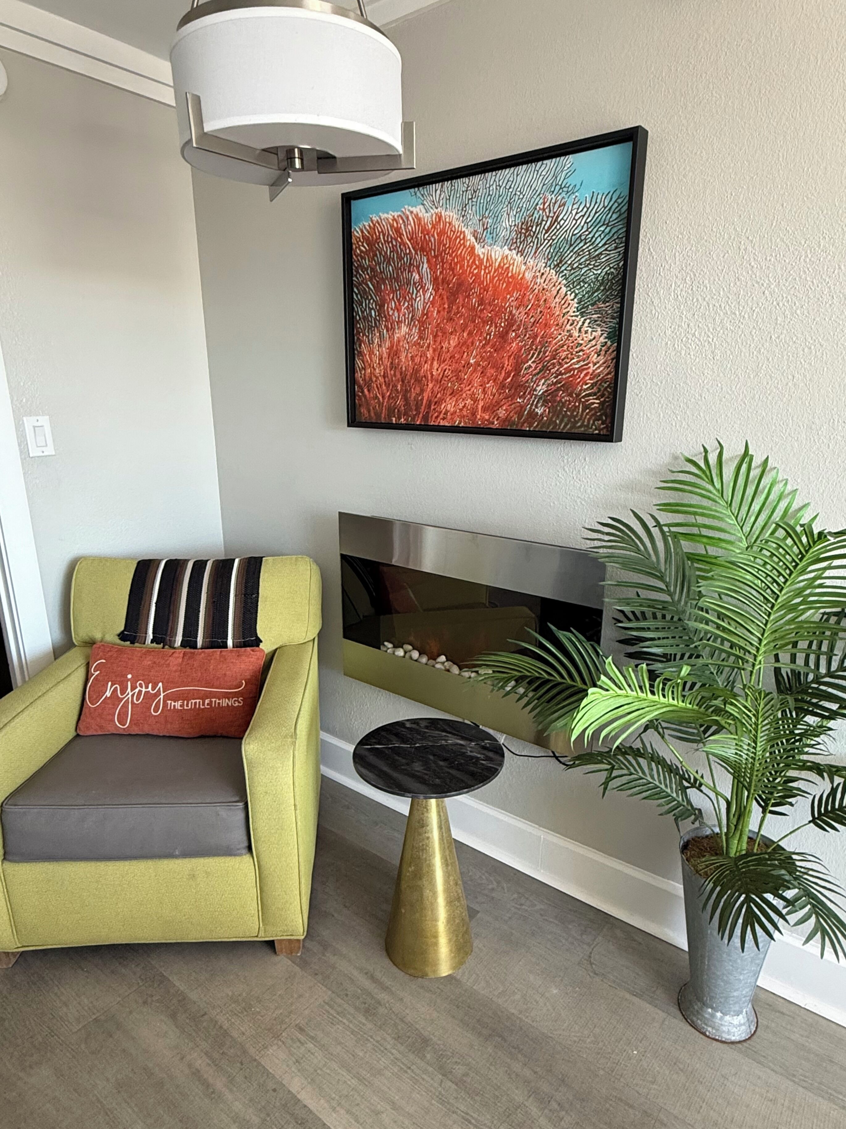 A living room with a green armchair, a small round table, a potted plant, a fireplace, and a framed picture on the wall.