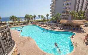 Ein Schwimmbecken mit Liegestühlen und Sonnenschirmen, im Hintergrund ein Strand und ein Hotelgebäude.