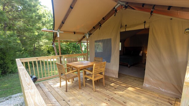 A wooden deck with a table and chairs, a covered area with a sleeping area inside, and a view of trees. - Settle Into a Cozy Lakeside Glamping Escape (Onalaska)