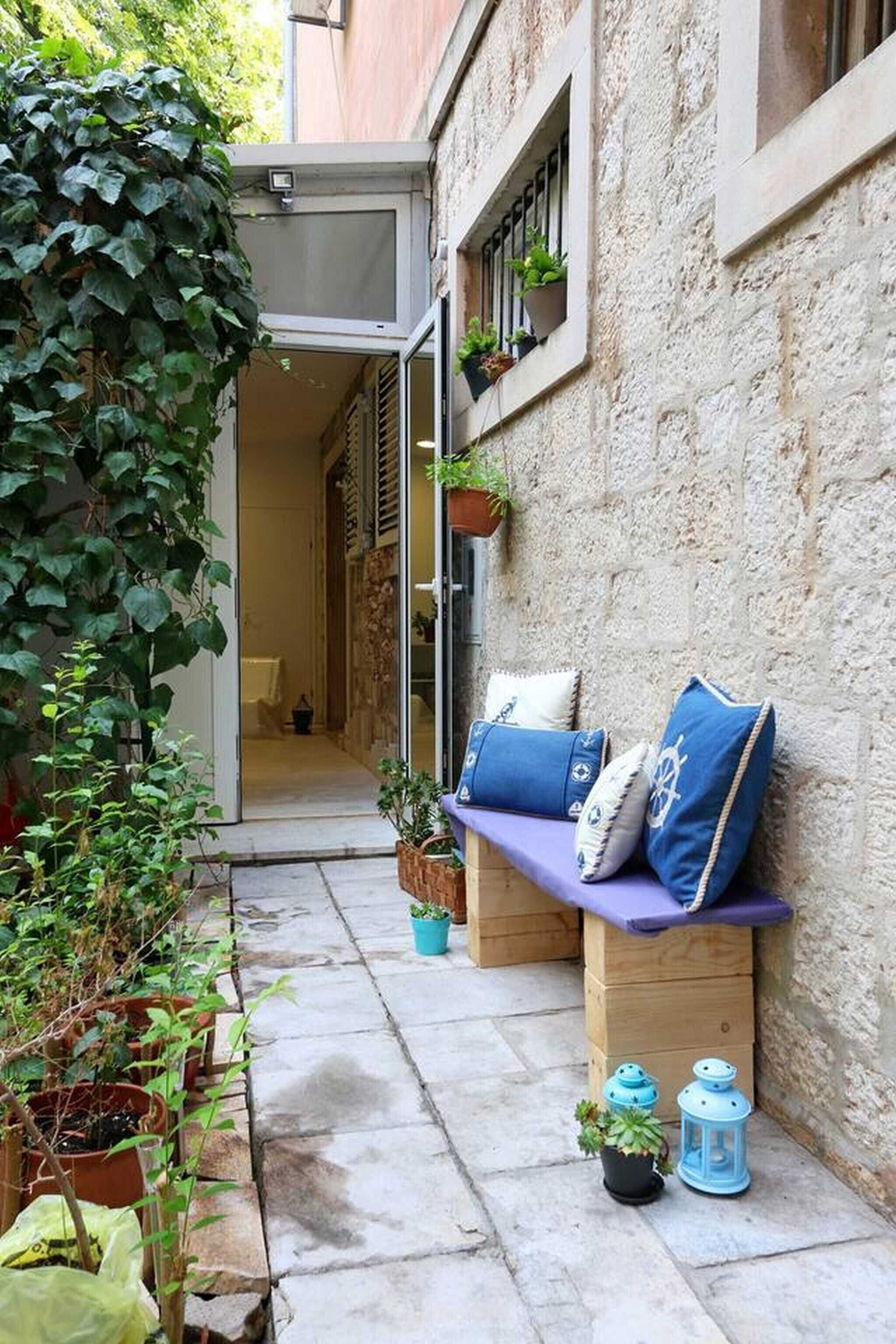 A narrow outdoor corridor with a bench, potted plants, and a door leading to an interior space.