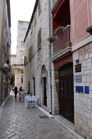Une rue étroite bordée d’immeubles en pierre, avec des terrasses et des enseignes indiquant la présence d’un restaurant et d’un bar.