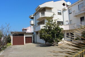 Un edificio residencial de varios pisos con un camino pavimentado, una puerta de garaje marrón y un pequeño árbol en primer plano.