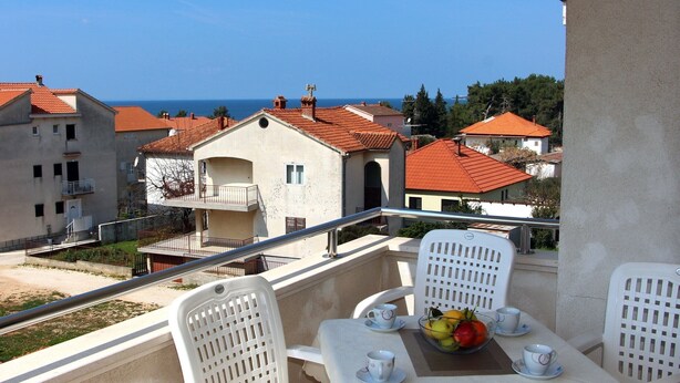 A balcony with white plastic chairs and a table, overlooking a residential area with red-tiled roofs and a view of the sea. - Zadar Coastal Haven - One Bedroom Apartment With Sea View (Zadar)