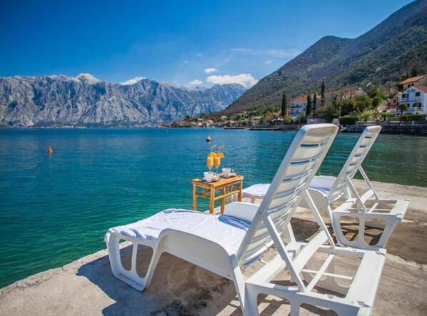 A lakeside view with two white lounge chairs and a small table with a drink, overlooking a mountain range.