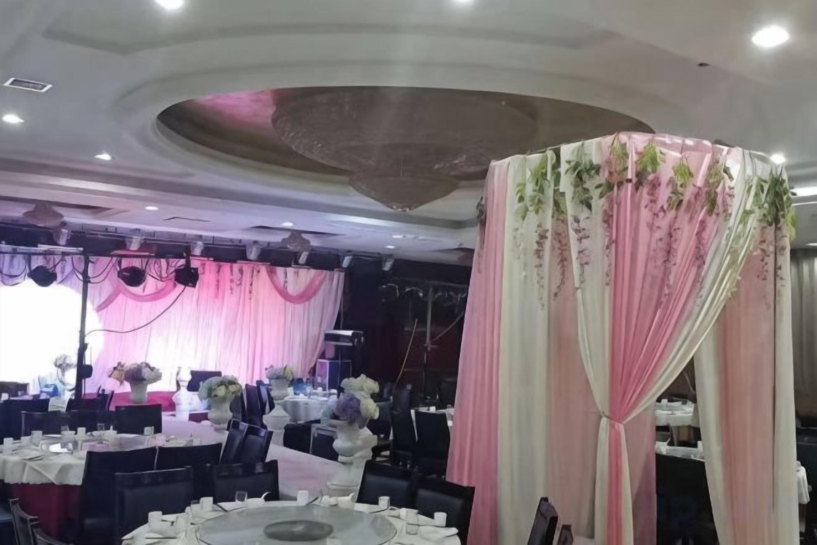 Salão de festas com mesas redondas, cortinas decorativas e arranjos florais.