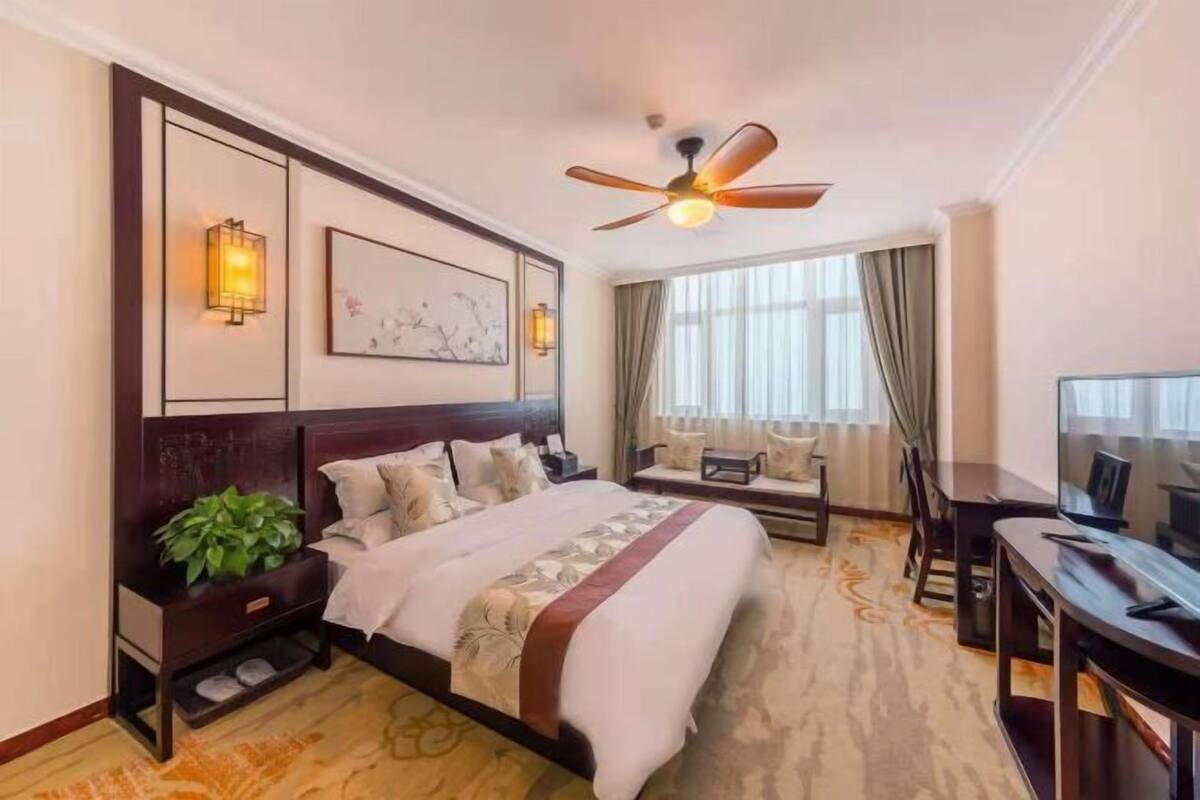 Ein Hotelzimmer mit einem großen Bett, einem Schreibtisch, einem Fernseher, einem Deckenventilator und einem Fenster mit Vorhängen.