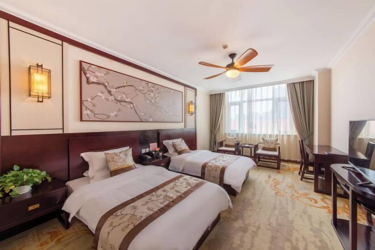 Ein Hotelzimmer mit zwei Betten, einem Deckenventilator, einem großen Wandbild, einem Fernseher und einem Essbereich.