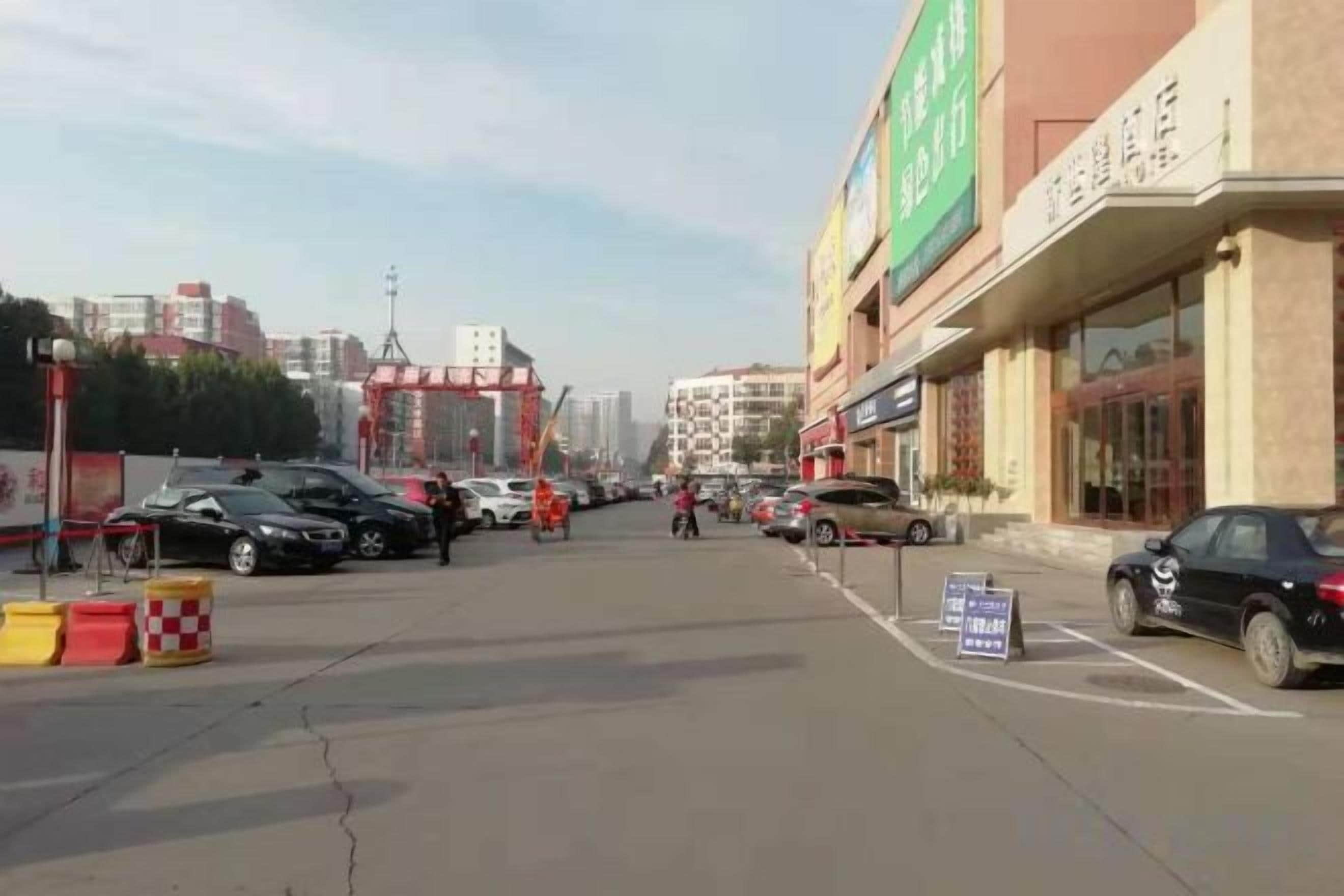 A street view with a building on the right, a parking lot with cars, and a red structure in the distance.