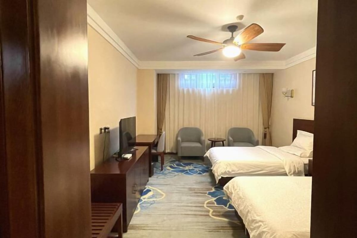 Ein Hotelzimmer mit einem Bett, zwei Sesseln, einem Schreibtisch, einem Fernseher und einem Deckenventilator.