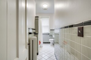 A narrow bathroom with white tiled walls and floor, a white toilet, and a shower area with a glass door. - Walk to Harvard Square: Apartment in Cambridge (Cambridge)