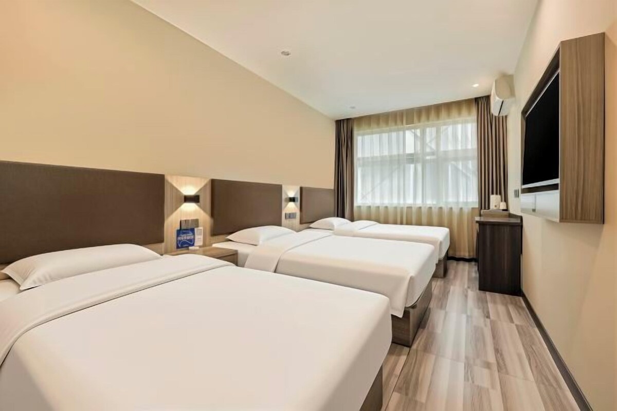 Habitación de hotel con tres camas, un televisor de pantalla plana, una mesita de noche con lámpara y una ventana con cortinas.