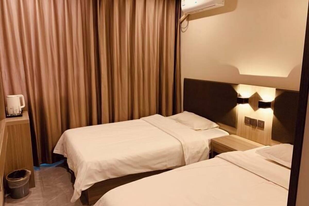 A hotel room with two beds, a bedside table with a lamp, a wall-mounted air conditioner, and a curtain.