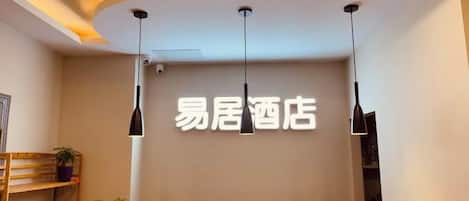 Zona de recepción de hotel con diseño moderno, que incluye un mostrador de recepción, iluminación colgante y un cartel en la pared.