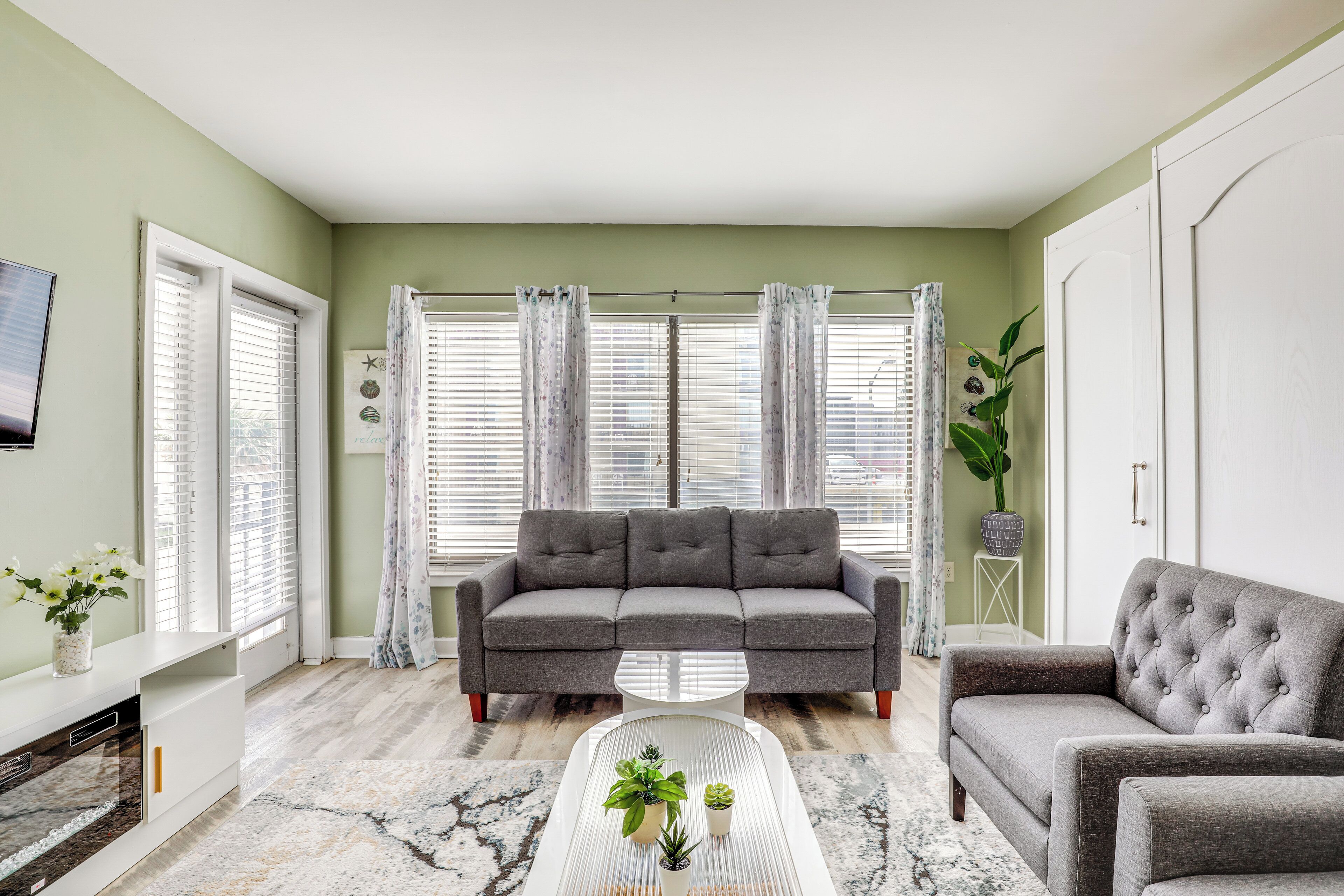 A living room with a grey sofa, a white coffee table with plants, and a large window with curtains.