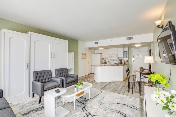 Ein modernes Wohnzimmer mit grauen Sofas, einem weißen Couchtisch und einer Küchenzeile mit weißen Schränken und einer Mikrowelle.