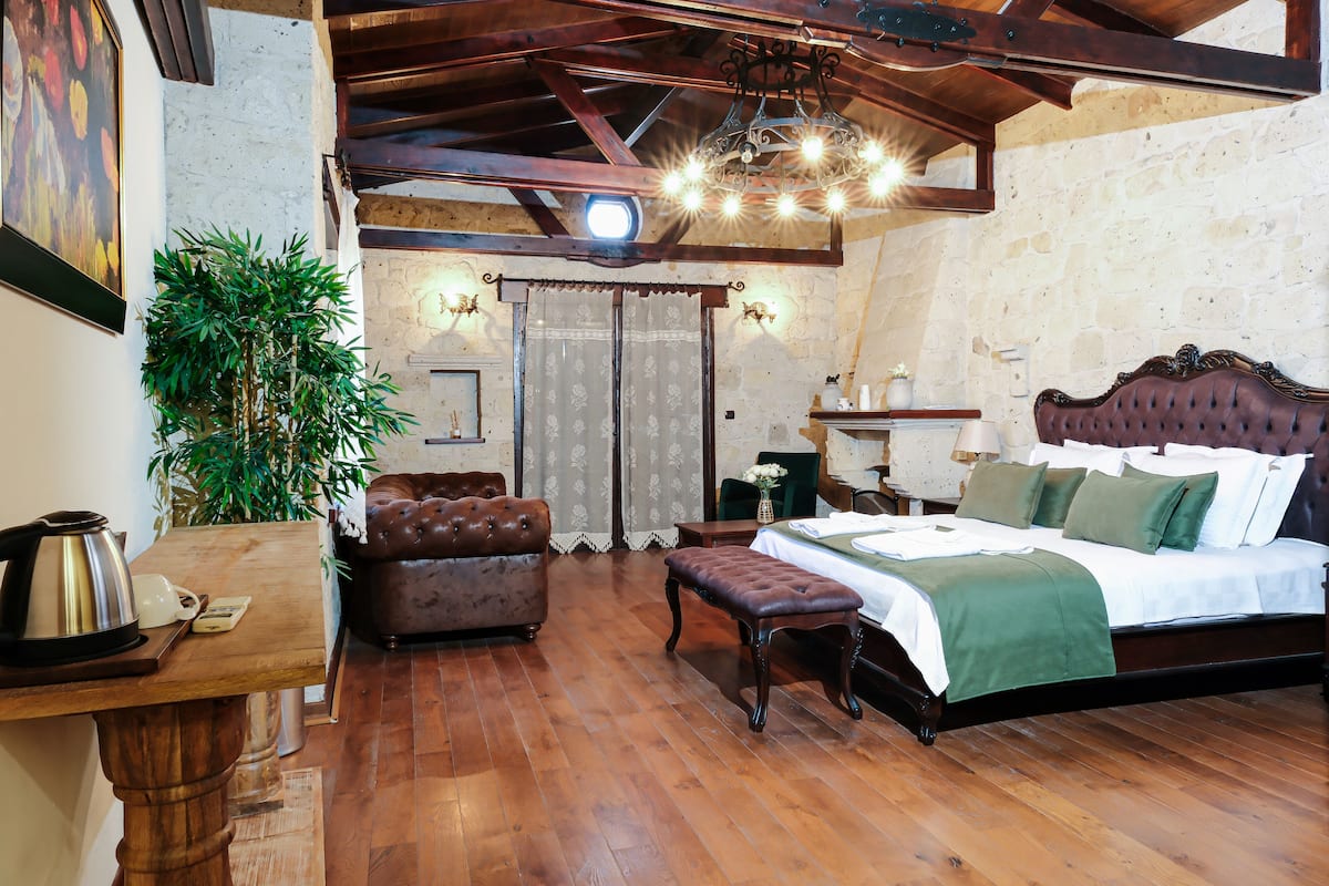 A bedroom with a wooden bed, a leather armchair, a wooden table with a kettle, and a large plant.
