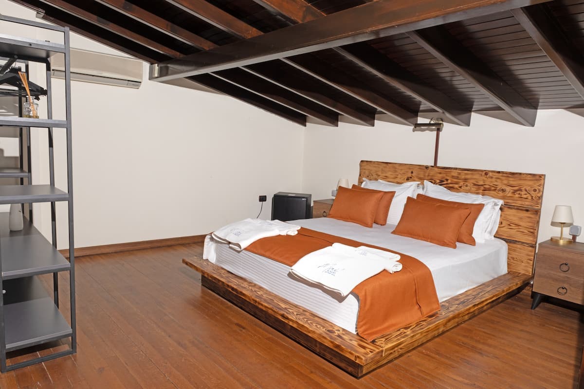 A bedroom with a wooden bed frame, white bedding, and an orange throw blanket. There is a nightstand with a lamp and a shelf with a speaker.