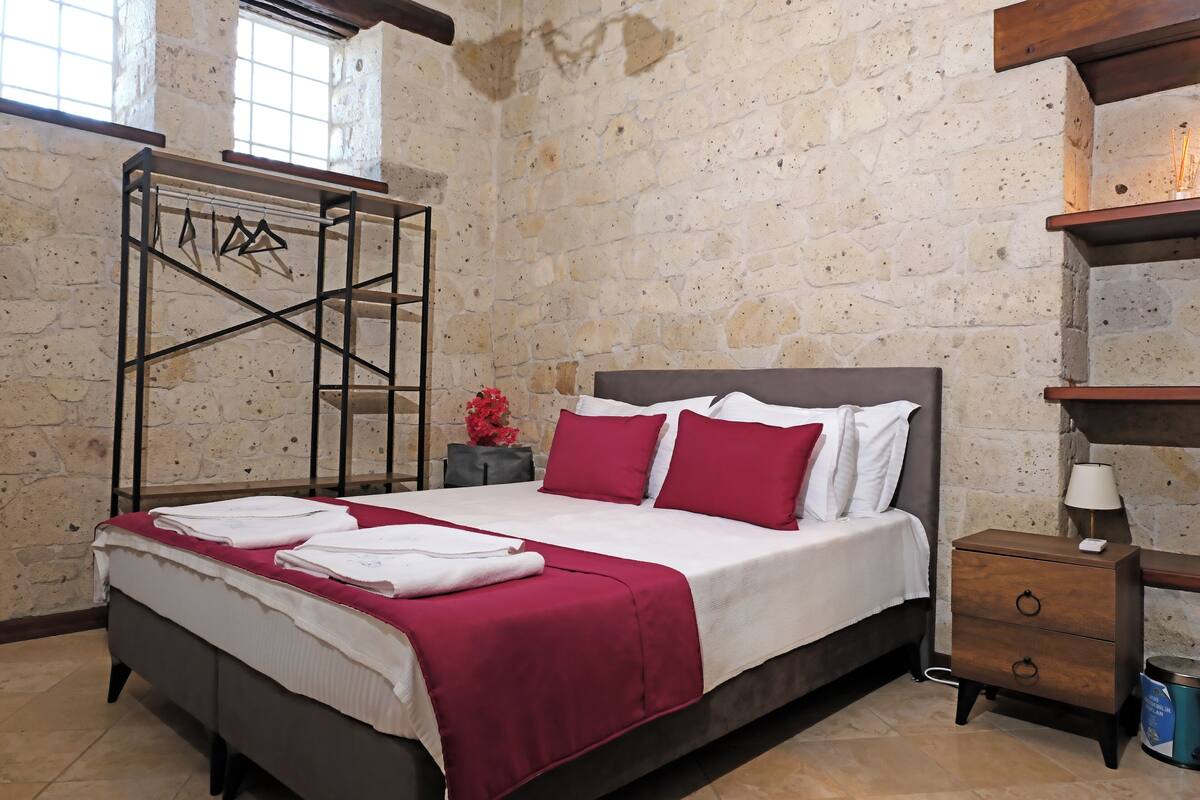 A bedroom with a stone wall, a bed with white and red bedding, a wooden nightstand, and a hanging clothes rack.