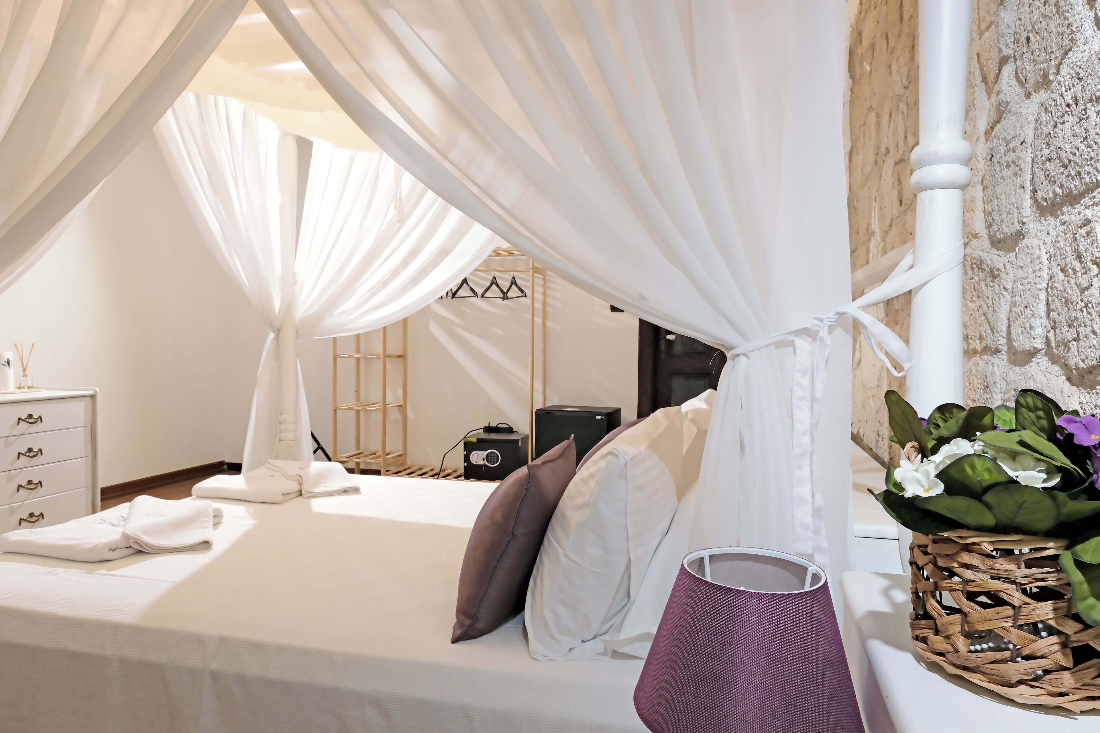 Una cama bien tendida con sábanas blancas, una mesita de noche con una lámpara y una cesta tejida con flores.