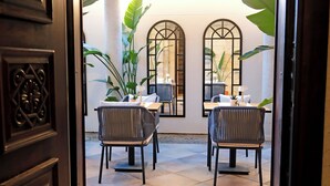 Une porte ouverte donne sur un espace repas comprenant deux tables, des chaises et de grandes plantes.