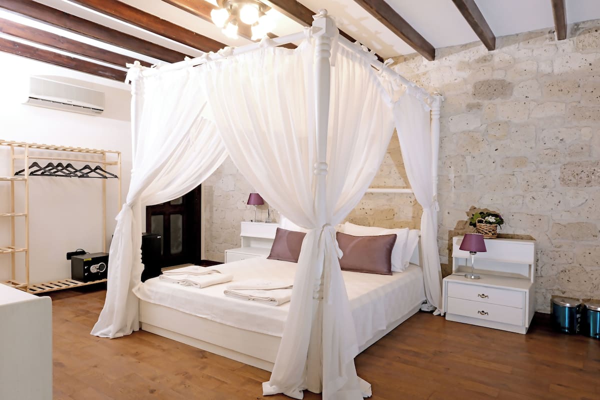 A bedroom with a four-poster canopy bed, wooden flooring, a white dresser, and a nightstand with a lamp.