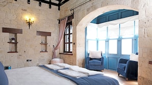 Une chambre à coucher avec un grand lit, deux fauteuils, un mur en pierre, une fenêtre avec des rideaux et un lustre.