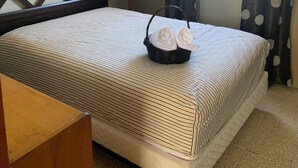 Una cama bien hecha con una colcha a rayas, una pequeña cesta de mimbre con toallas, un cuadro enmarcado en la pared y una ventana con cortinas.