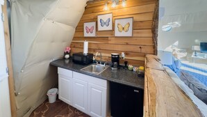 Een compacte keuken met een spoelbak, magnetron en koffiezetapparaat. Er hangen foto’s van vlinders in lijsten aan de muur en er staat een kleine prullenbak.