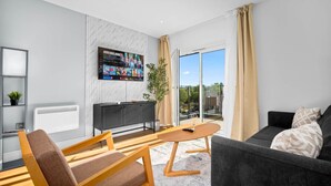 Un salon moderne doté d’un téléviseur à écran plat, d’un canapé, d’un fauteuil, d’une petite table et d’une porte-fenêtre coulissante donnant sur un balcon.