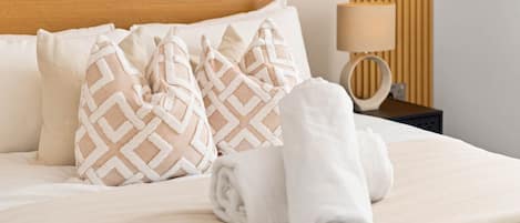 A neatly made bed with patterned pillows, a rolled towel, and a bedside lamp on a wooden nightstand.