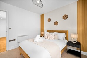 Une chambre d’hôtel moderne avec une tête de lit en bois, un lit bien fait avec du linge de lit blanc, une table de chevet avec une lampe et des œuvres d’art géométriques accrochées au mur.