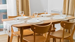 Una mesa de comedor dispuesta con platos blancos, vasos y un posavasos, rodeada de sillas de madera en una habitación con ventana y cortinas.