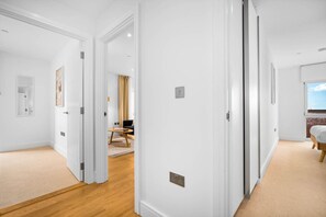 Un couloir aux murs blancs, doté d’un plancher en bois et d’une porte donnant sur une pièce où se trouvent un lit et une fenêtre.