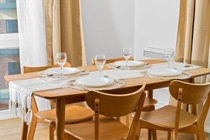 A dining table set with white plates, glasses, and a placemat, surrounded by wooden chairs in a room with a window and curtains.
